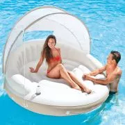 Intex Canopy Island - A smiling woman in a red bikini sits on a large, round inflatable pool float with a beige base and a white detachable canopy, while a man is partially visible in the water beside it.